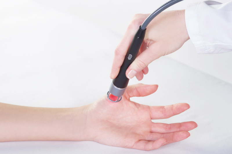 A healthcare professional applies a handheld medical device to a patient's wrist.